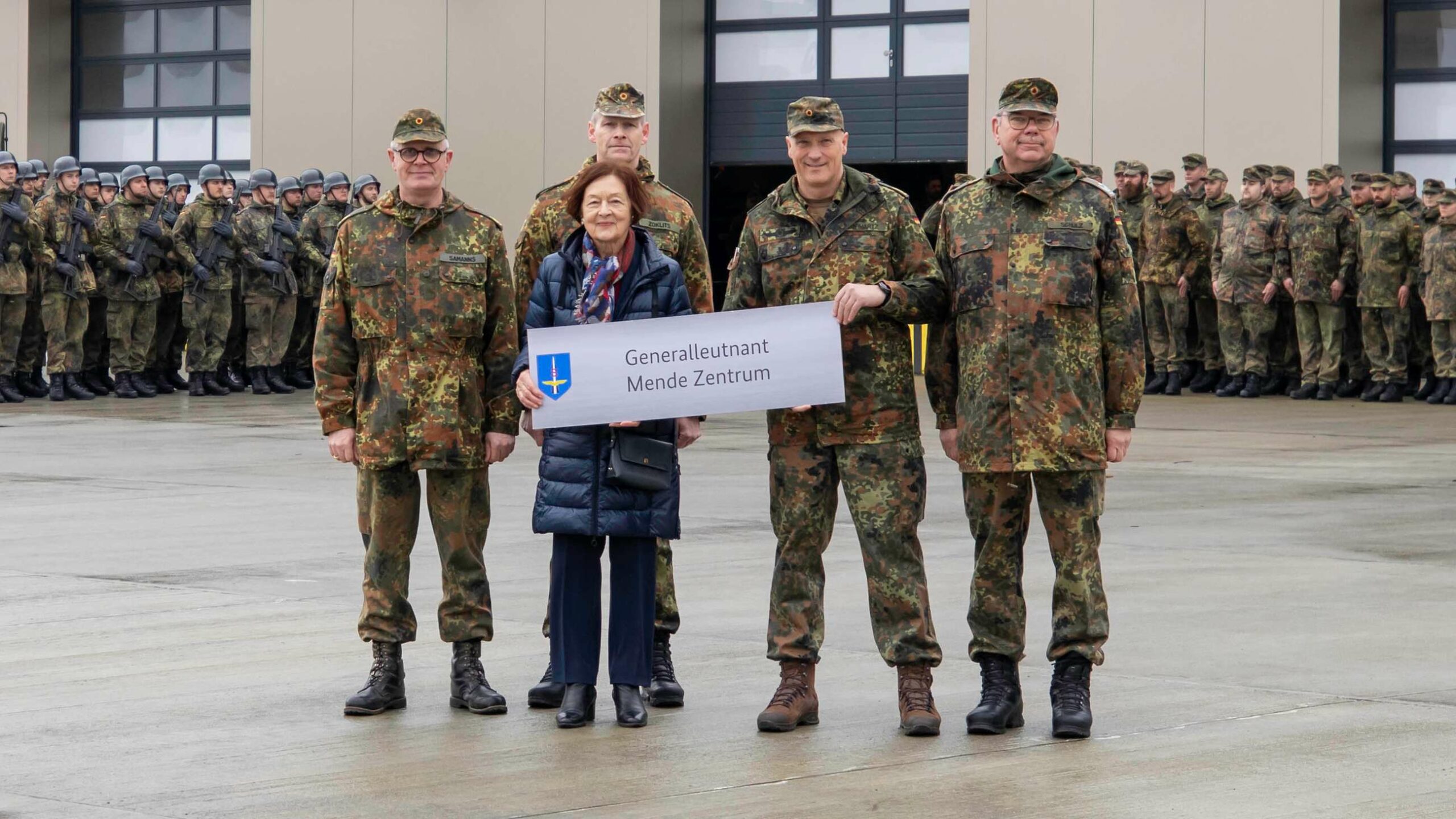 Generalleutnant Ingo Gerhartz (2. v. r.) und Karin Mende, die Ehefrau des verstorbenen Namenspatrons, übergeben bei einem feierlichen Appell das neue Gebäudeschild an die Führungskräfte der bodengebundenen Luftverteidigung auf dem Platz vor dem Neubau des Ausbildungszentrums Flugabwehrraketen.
